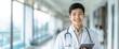 © zakiroff - Smiling young male doctor wearing white coat with stethoscope, holding tablet, standing in bright hospital corridor. Banner with copy space.