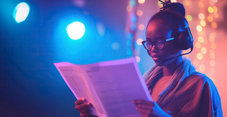 Black young female event manager or party planner woman reading documen while wearing headset earphones with microphone, live concert party show in background with colorful spotlights and copy space