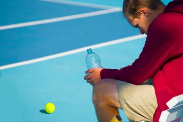 Wall Mural - A young girl tennis player is resting while sitting on a bench in between games and drinks water	