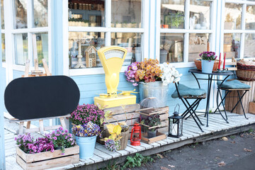 Wall Mural - Facade blue сozy home with garden furniture. Farmhouse with wicker baskets and flowers on terrace. Wooden table and chair on veranda. Showcase with chrysanthemums, scales and plants in a flower shop