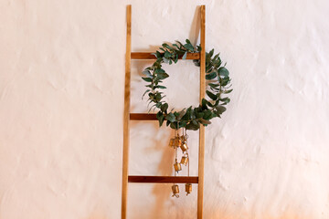 Wall Mural - Wooden ladder against a white wall. Boxwood wreath hanging on an old wooden stepladder. Green wreaths with leaves. Wreath made of branches with vintage bells. Green olive wreath. Festive Xmas decor