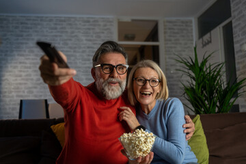 Poster - Couple Watches TV together while Sitting on a Couch in the Living Room. TV Programming Concept. Relaxed Senior Couple Watching Television Together Sitting On Sofa And Pointing Remote Controller