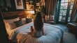 © Artsaba Family - Back view of a woman sitting on a luxurious bed, gazing out of a window to an urban scene.