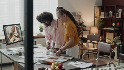 Medium shot of two ethnically diverse interior designers standing by office desk with blueprints on it and 3d rendering software on desktop computer, having brainstorm discussion of ideas