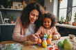 Easter Family traditions. Loving ethnic young mother teaching happy little kid soon to dye and decorate eggs with paints for Easter holidays while sitting together at kitchen table