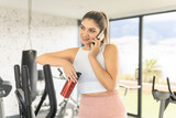 Fototapeta Przestrzenne - young woman taking a break talking on cell phone and drinking water in the gym