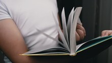 Close Up, Unrecognizable Boy Flipping Through Pages Of A Book. Person Standing, Holding A Book In Hands, Quickly Turning Pages In Slow Motion In Room. Reading. Blurred Background. Concept Education.