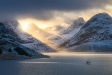 Snowy mountains in low clouds, bright sunbeams, boat in sea bay at colorful sunset in winter. Lofoten islands, Norway. Amazing landscape with rocks in snow, golden sun rays. North seaside. Nature