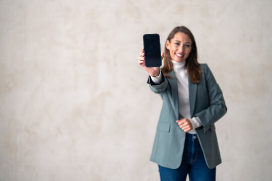 Smiling female showing mobile smartphone display, marketing or app on mockup. Studio portrait of a charming young woman holding a cellphone with a blank screen against a gray background.