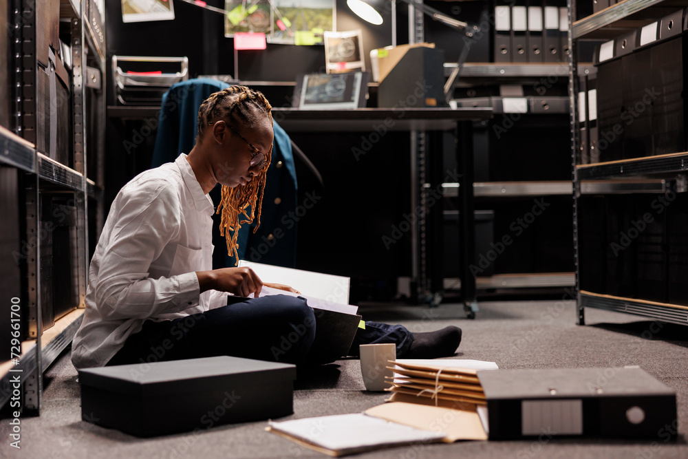 Law enforcement agent reading crime case file while sitting on floor in office. African american woman investigator analyzing forensic report and archival records at night time