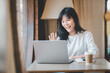 © Satori Studio - Business freelance concept, Cheerful young woman engaging in a video call on her laptop in a cozy cafe setting.