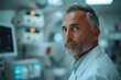 © P - A serious man in a white medical coat stands against an operating room backdrop with medical equipment.