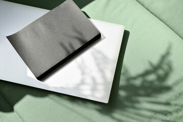 Closed laptop computer and black notebook with empty cover on sage green chair textile background with abstract natural floral sunlight shadows, home office workspace