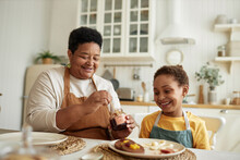 Cheerful African American Grandma Serving Breakfast For Her Granddaughter Adding Jam To Pancakes On Plate