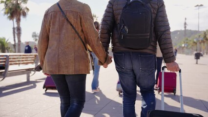 Wall Mural - Unrecognizable adult tourist man and woman holding hands backwards while walking down the street with suitcase on vacation. Close-up of the bodies of two people from behind