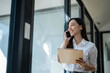 © Wasana - Smiling young Asian businesswoman having a phone conversation while holding a clipboard in a well-lit modern office setting..