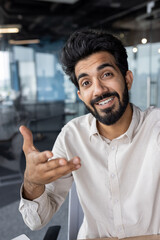 Wall Mural - Vertical close-up photo of a young Indian man working in the office and talking to the camera on a video call, smiling and gesturing