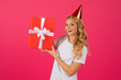 Leinwandbild Motiv Happy blonde woman wearing birthday hat holding gift, pink background
