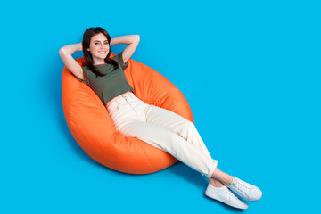 Poster - Full size photo of satisfied girlish woman wear green t-shirt sit on pouf hold palms behind head chill isolated on blue color background