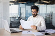 © Tetiana - Focused indian businessman analyzing documents at office desk with laptop and smartphone