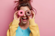 Young woman is smiling while she is watching through donuts on her eye. Donat is with pink chocolate dipping and colorful crumbles