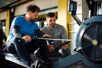 Wall Mural - Young coach and mature athlete going through exercise plan during workout on rowing machine in gym.