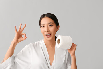 Poster - Young Asian woman in gown with toilet paper showing OK on light background