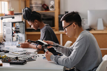 Young technician sitting at table and fixing faulty drone, his black colleague working on background