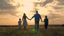 Happy family walks on green grass in meadow in spring. Parents, children are walking in park at sunset. Family, child, walk through summer field holding hands. Dad mom son daughter together, nature
