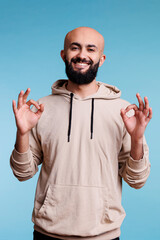 Wall Mural - Smiling arab man making ok sign with fingers and looking at camera with cheerful facial expression. Carefree person showing okay approval gesture with two hands studio portrait