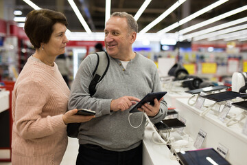 Wall Mural - Mature man and woman standing in salesroom of electronic shop and choosing tablet.