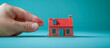 © Gejsi - Hand holding a brick building a miniature house over blue background