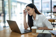 © Wasana - A stressed young Asian businesswoman massages temples while focusing on a laptop screen in a well-lit office environment..