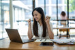 © Wasana - A friendly Asian businesswoman waves hello during a video call on her laptop, holding a coffee cup in a bright office setting..