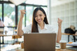 © Wasana - A delighted Asian businesswoman raises her fists in victory while looking at her laptop in a well-lit office space..