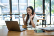 © Wasana - A friendly Asian businesswoman waves hello during a video call on her laptop, holding a coffee cup in a bright office setting..