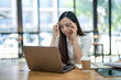© Wasana - A stressed young Asian businesswoman massages temples while focusing on a laptop screen in a well-lit office environment..