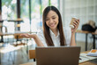 © Wasana - A friendly Asian businesswoman waves hello during a video call on her laptop, holding a coffee cup in a bright office setting..