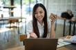 © Wasana - A friendly Asian businesswoman waves hello during a video call on her laptop, holding a coffee cup in a bright office setting..