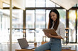 © Wasana - An energetic Asian businesswoman is actively working with a laptop while referencing notes on a clipboard in a vibrant office space..