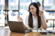© Wasana - A friendly Asian businesswoman waves hello during a video call on her laptop, holding a coffee cup in a bright office setting..