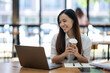 © Wasana - A friendly Asian businesswoman waves hello during a video call on her laptop, holding a coffee cup in a bright office setting..
