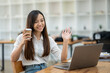 © Wasana - A friendly Asian businesswoman waves hello during a video call on her laptop, holding a coffee cup in a bright office setting..