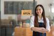 © Wasana - A confident and smiling Asian female cafe owner stands proudly with crossed arms behind a 'Welcome Open' sign at her coffee shop..