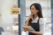 © Wasana - Smiling Asian cafe owner writes in her notebook, ready to welcome customers, with a 'Welcome Open' sign hanging in the background..