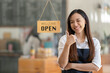 © Wasana - Asian female cafe owner with a welcoming smile presents the open sign at her shop, signaling readiness for customers..