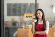© Wasana - Asian female cafe owner with a welcoming smile presents the open sign at her shop, signaling readiness for customers..