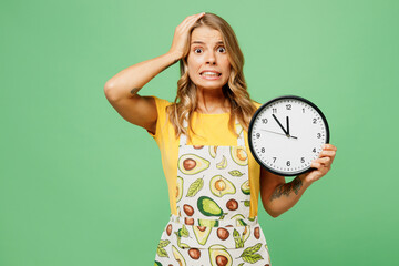 Young disappointed sad housewife housekeeper chef cook baker woman she wear apron yellow t-shirt hold head clock show time look camera isolated on plain green background studio. Cooking food concept.