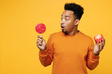 Young Man Wear Orange Sweatshirt Casual Clothes Hold In Hand Pink Lollipop Red Apple Isolated On Plain Yellow Background Studio Portrait. Proper Nutrition Healthy Fast Food Unhealthy Choice Concept.