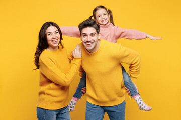 Poster - Young parents mom dad with child kid girl 7-8 years old wear pink knitted sweater casual clothes giving piggyback ride to joyful, sit on back isolated on plain yellow background. Family day concept.